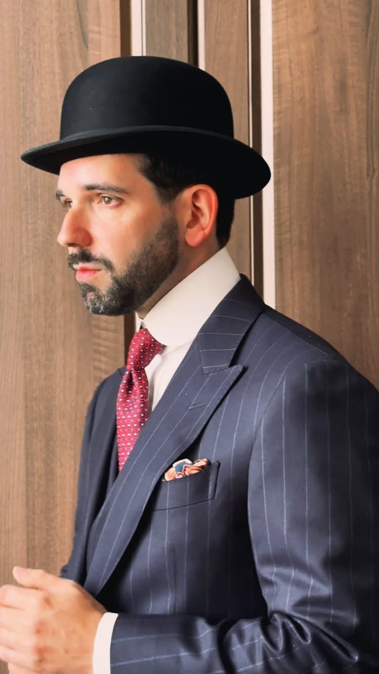 Man in profile wearing a black bowler hat, pinstriped suit, and red patterned necktie.