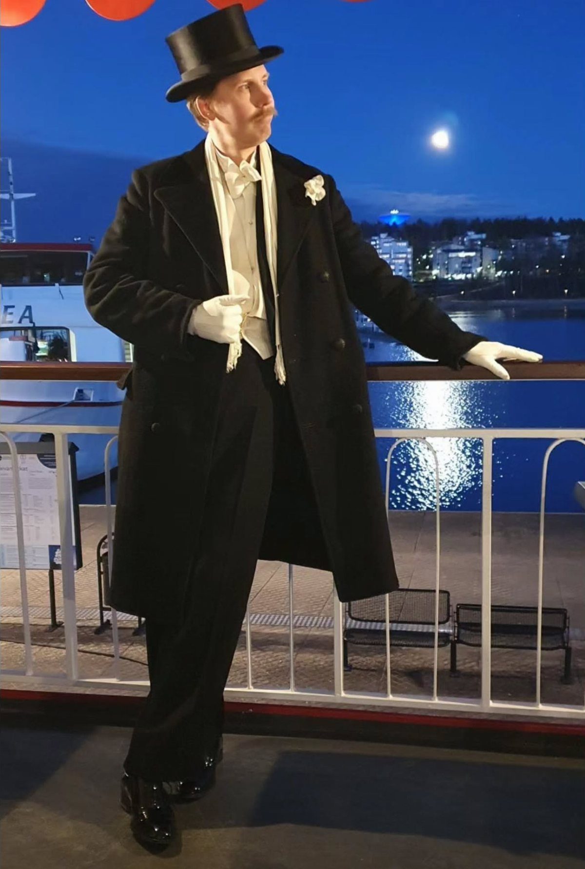 Man in formal 1880s attire wearing a long black coat and top hat on a boat.