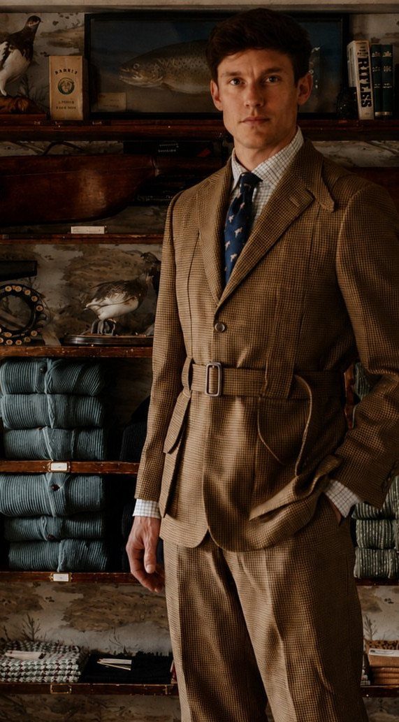 A man wearing a brown patterned Norfolk jacket with a belt and a blue necktie.