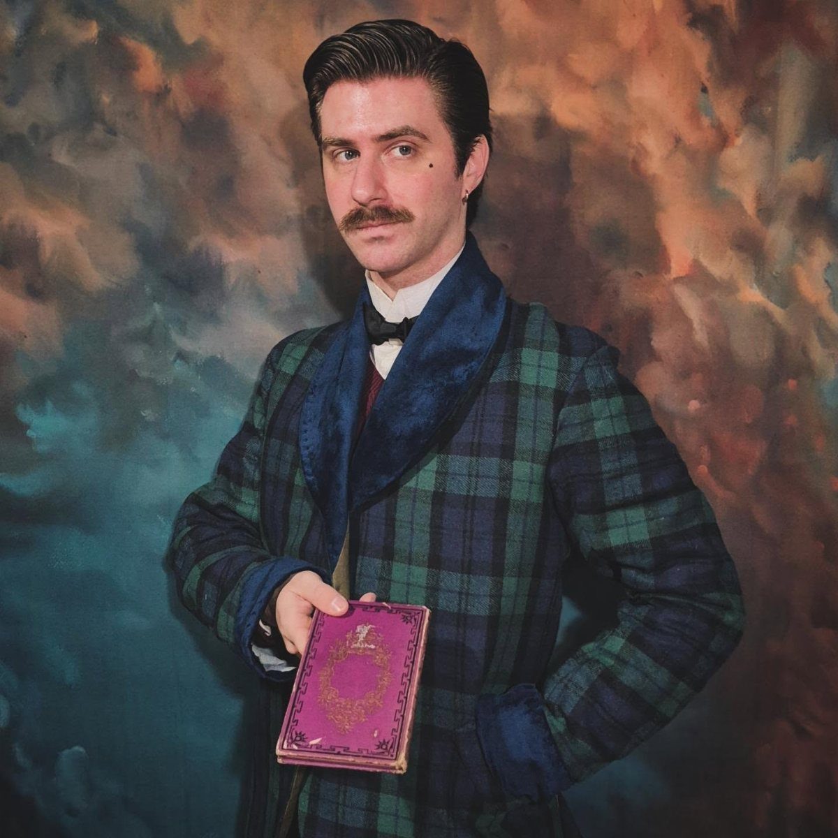 Man with a mustache wearing a plaid smoking jacket and bowtie holding a pink book.