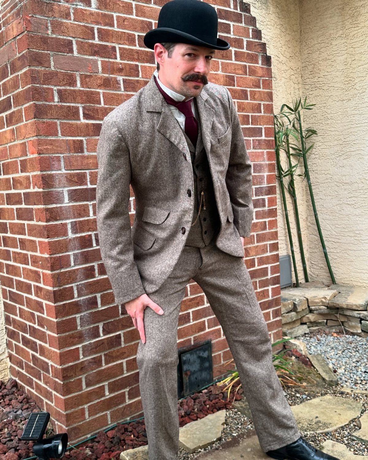 A man in a brown tweed three-piece suit and bowler hat posing by a brick wall.
