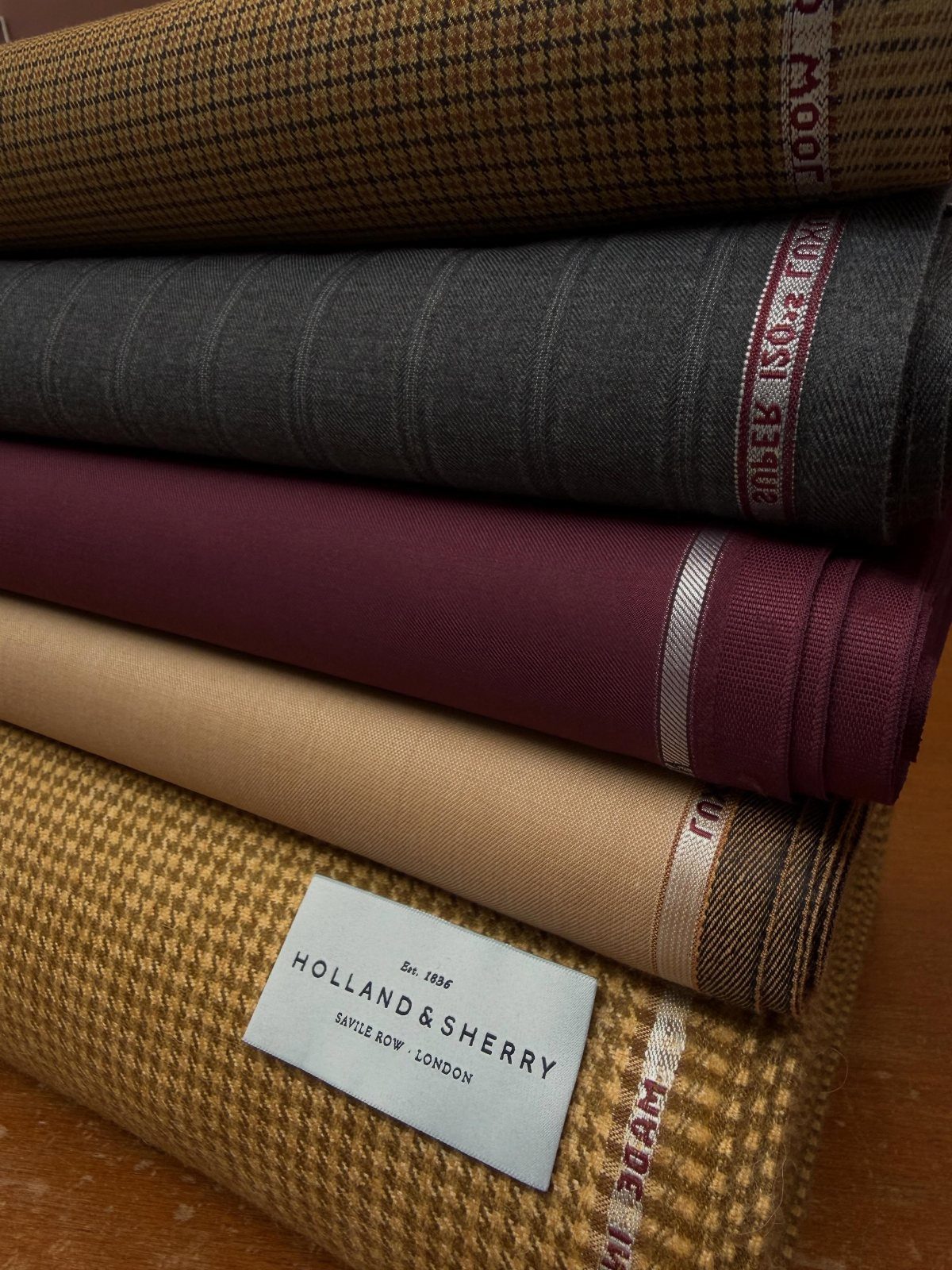 Stacked rolls of patterned and solid colored textile fabrics in brown, grey, burgundy, and tan.