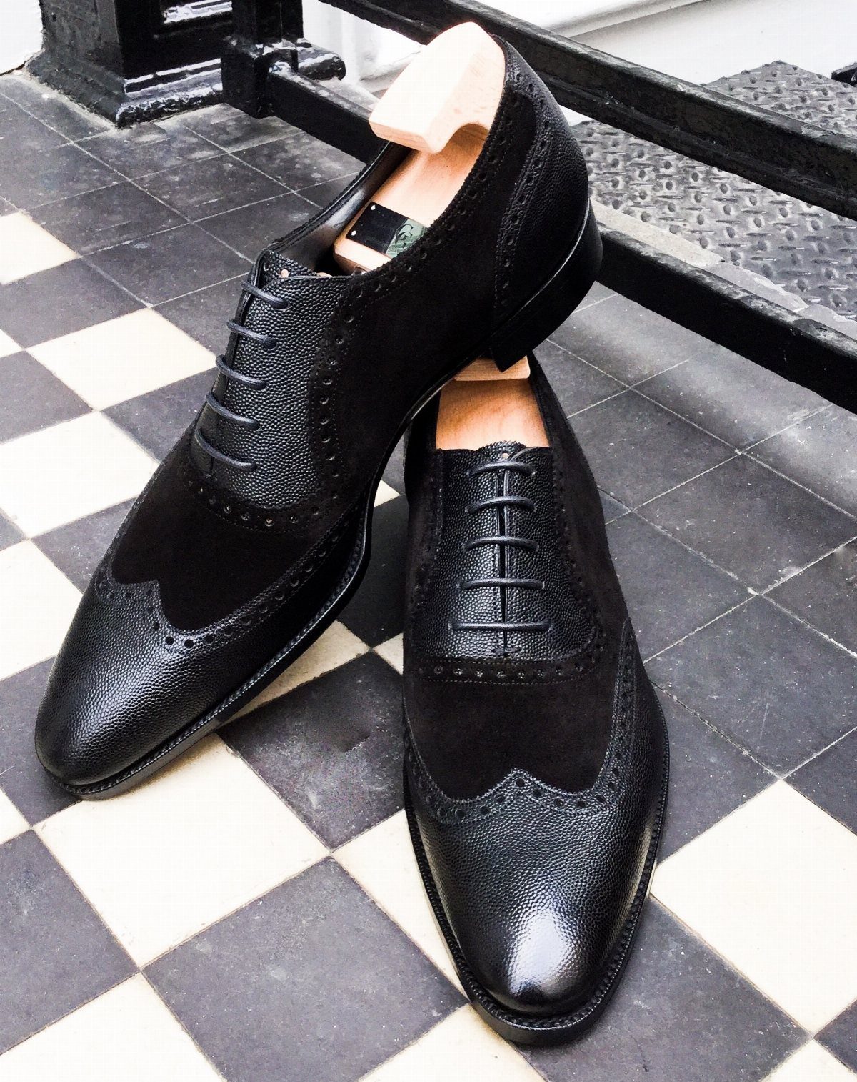 Black leather and suede wingtip Oxford shoes resting on a checkered floor.