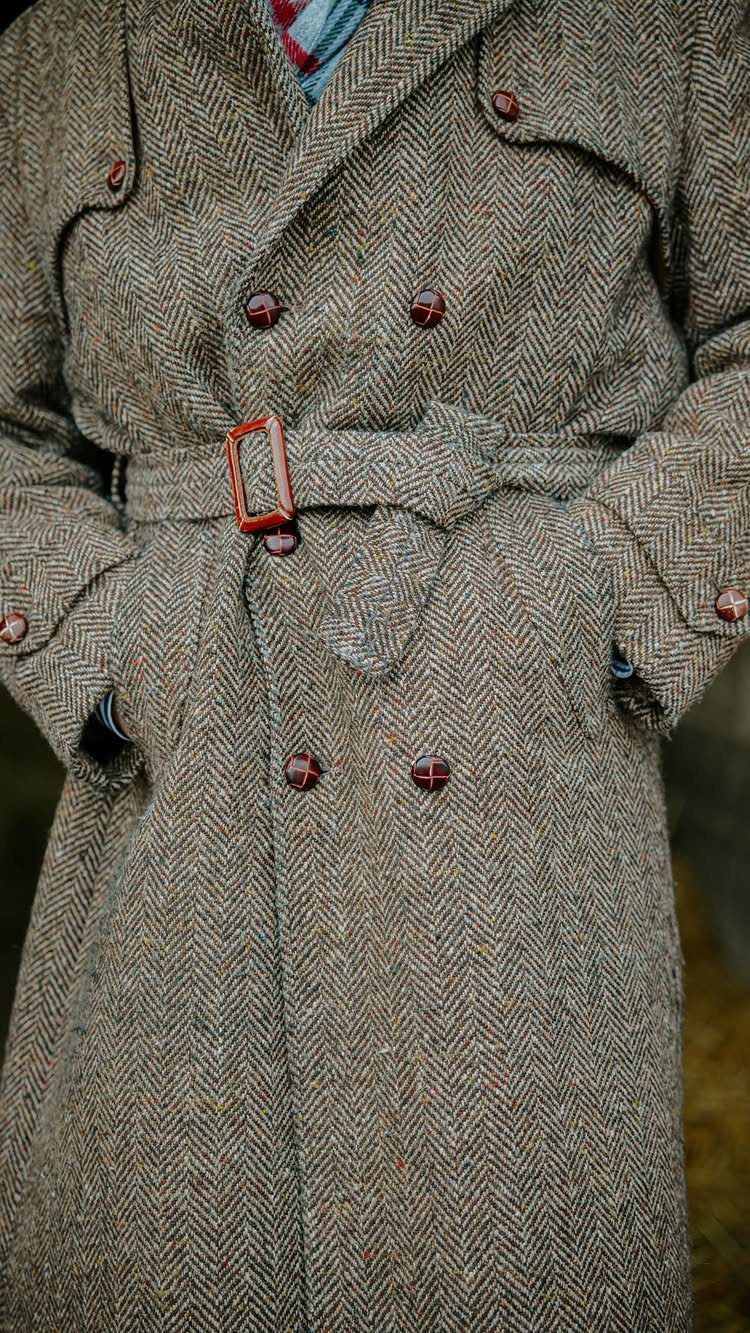 A person wearing a heavy brown herringbone pattern trench coat with a belted waist.