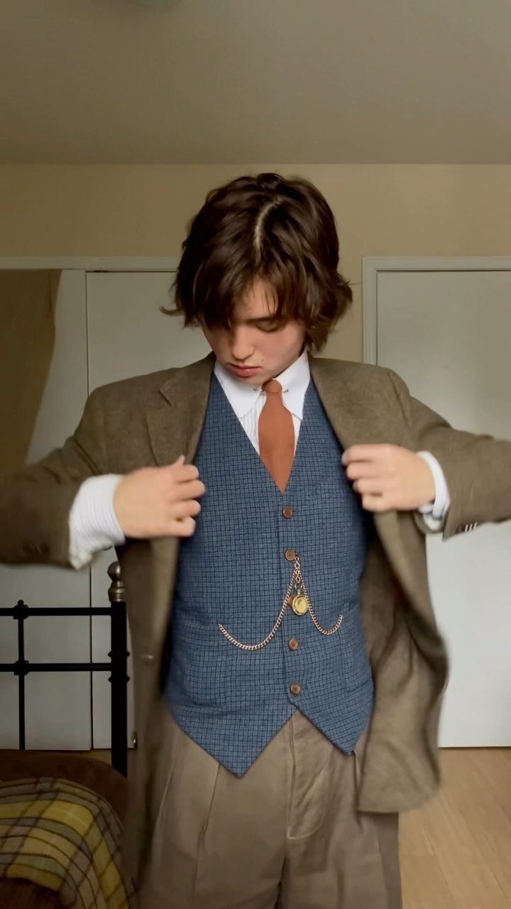 Young man posing in a brown blazer, blue waistcoat, orange tie, and tan trousers.