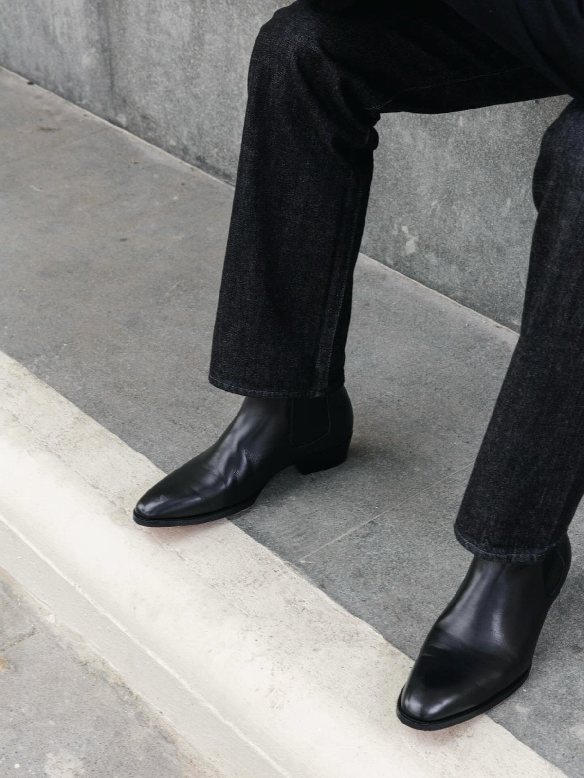 Black leather Chelsea boots worn with dark slim-fit trousers on a concrete step.