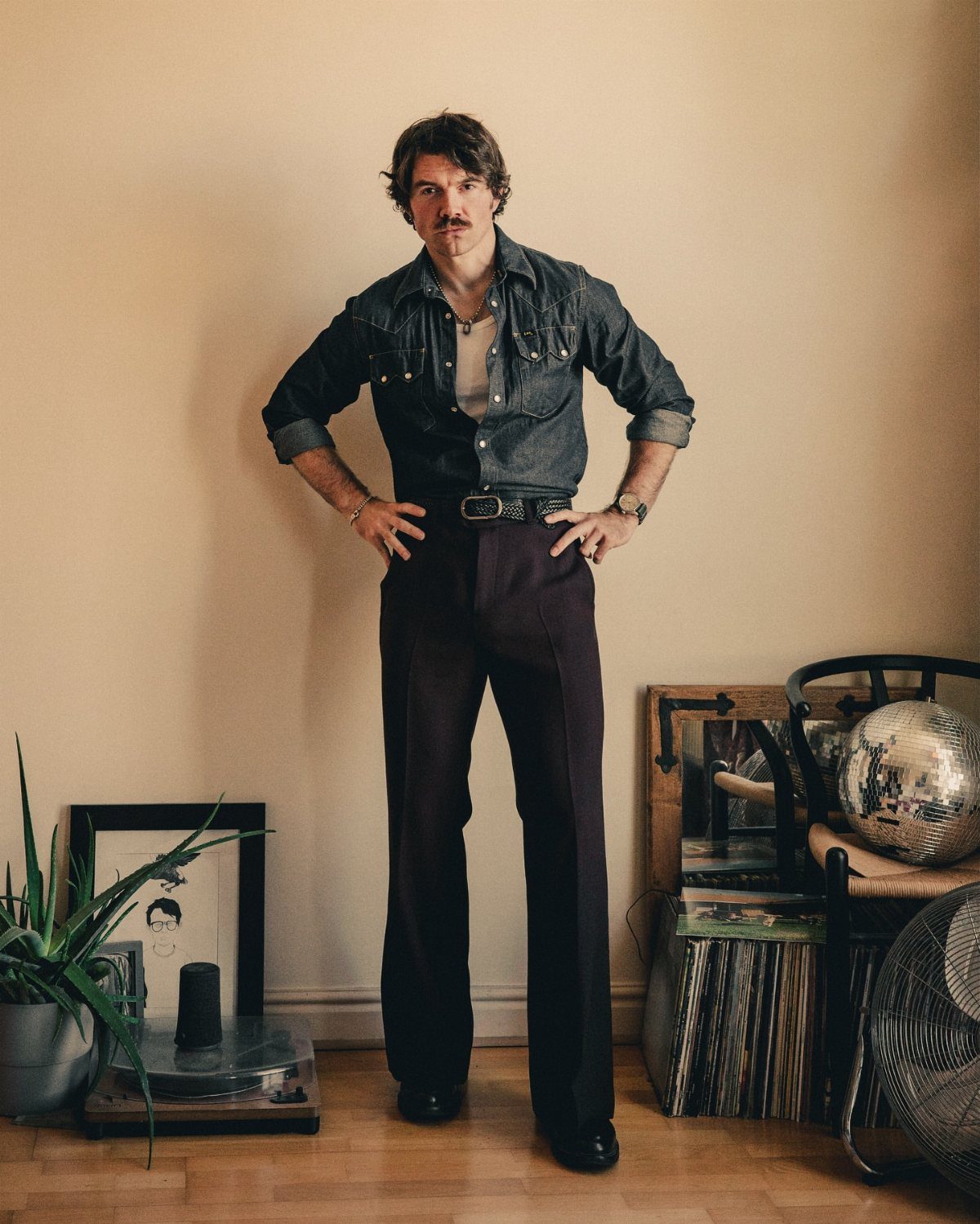 A man posing in a denim shirt and dark flared trousers against a plain wall.