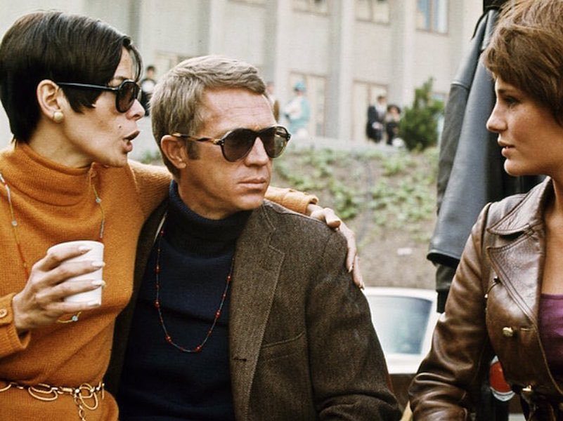 A man in dark sunglasses and a navy turtleneck sits between two women outdoors.