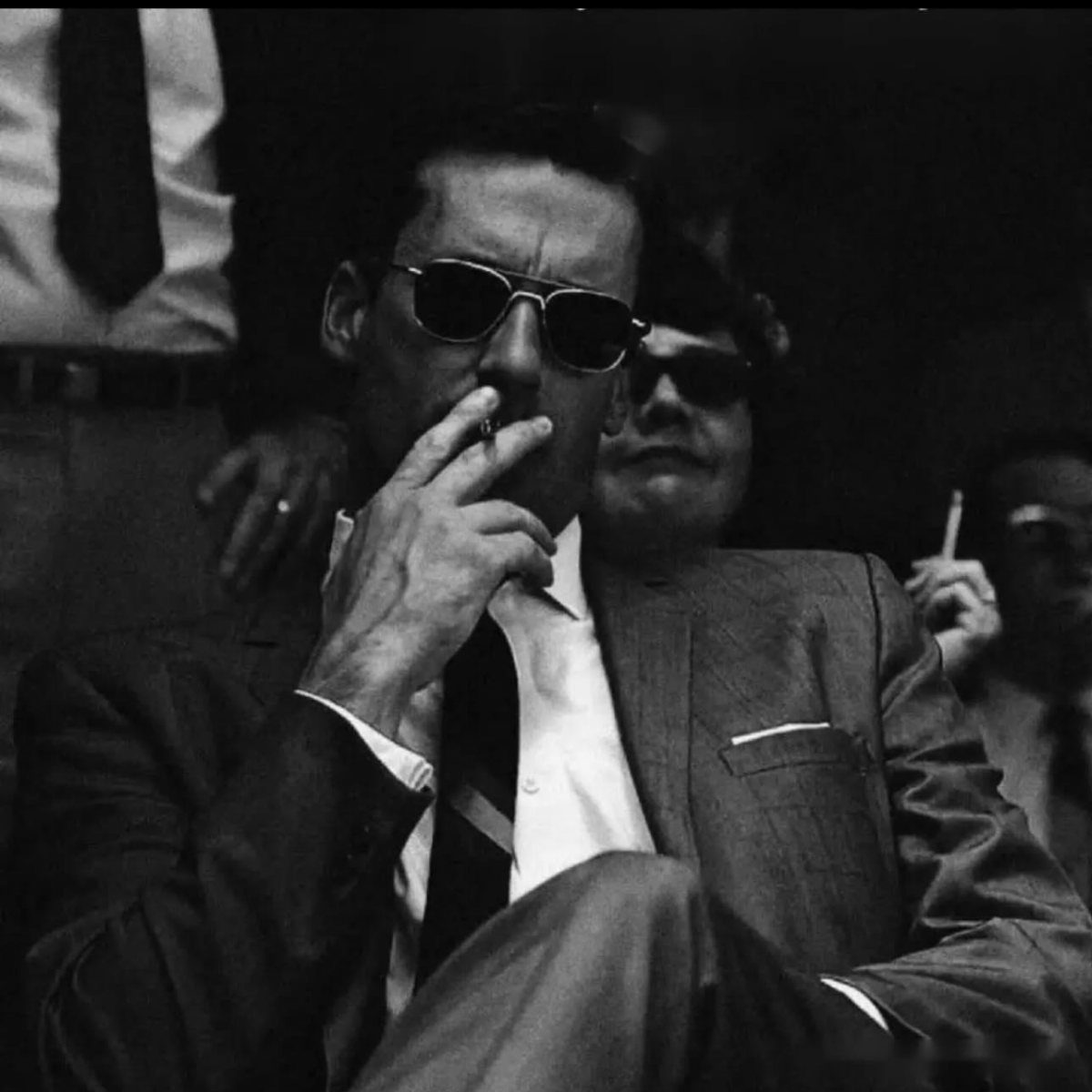 A man in a suit and sunglasses smoking a cigarette in a dark setting.