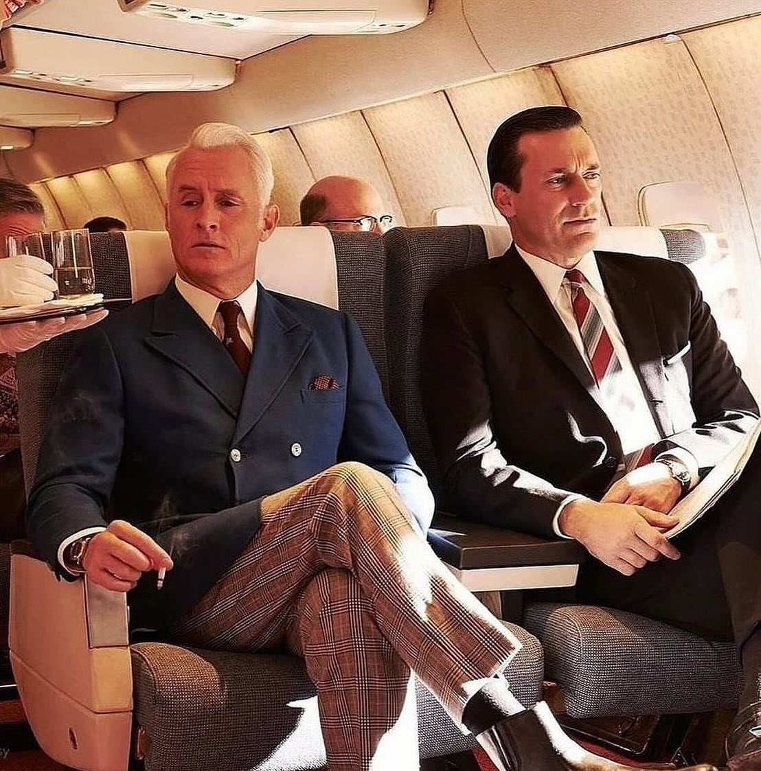 Two men in mid-century tailored suits sitting in airplane seats during a flight.