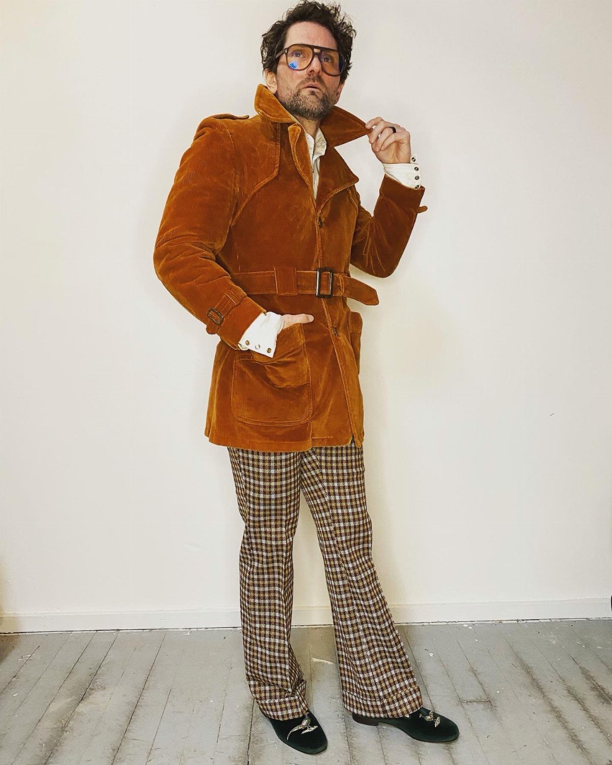 A man wearing a brown corduroy coat and patterned flared trousers poses against a white wall.