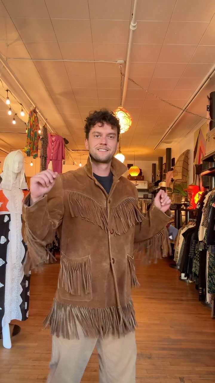 A man smiling while wearing a vintage brown suede fringe jacket in a clothing store.