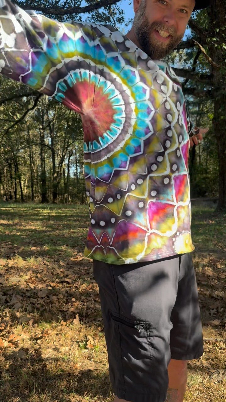 Man smiling in a colorful tie-dye t-shirt with a large mandala pattern outdoors.