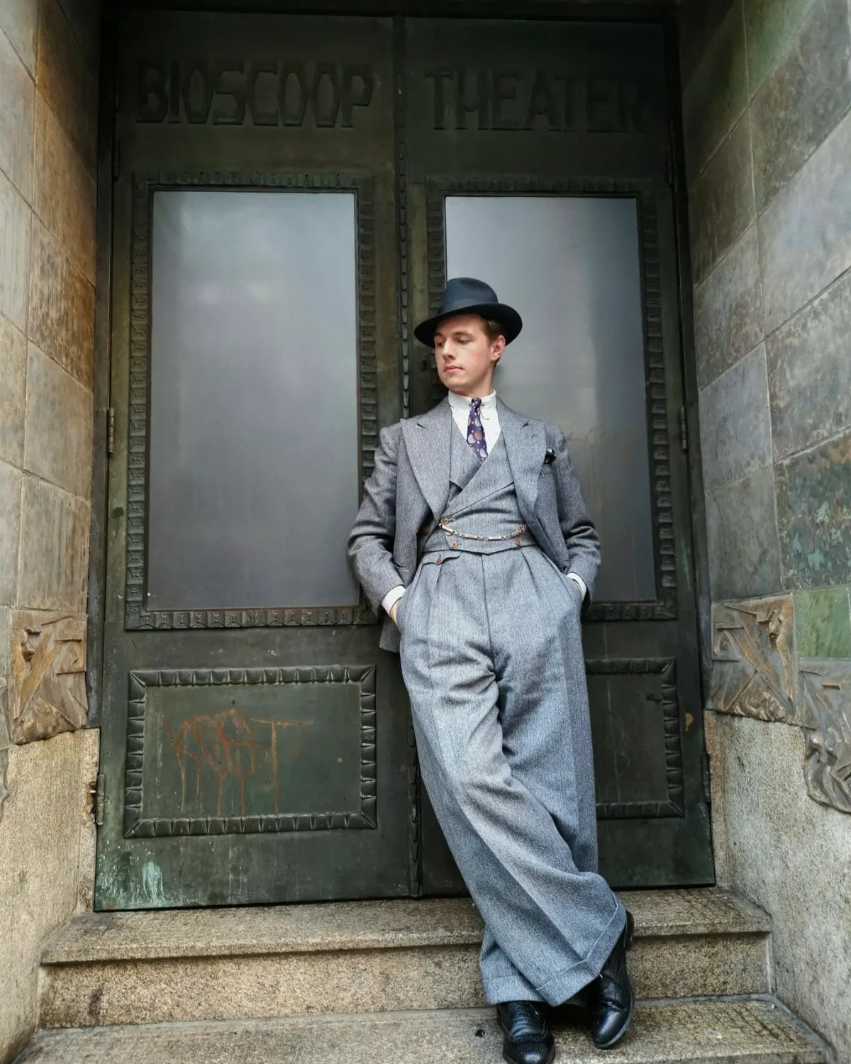Man in a grey 1930s three-piece suit and fedora leaning against theater doors.
