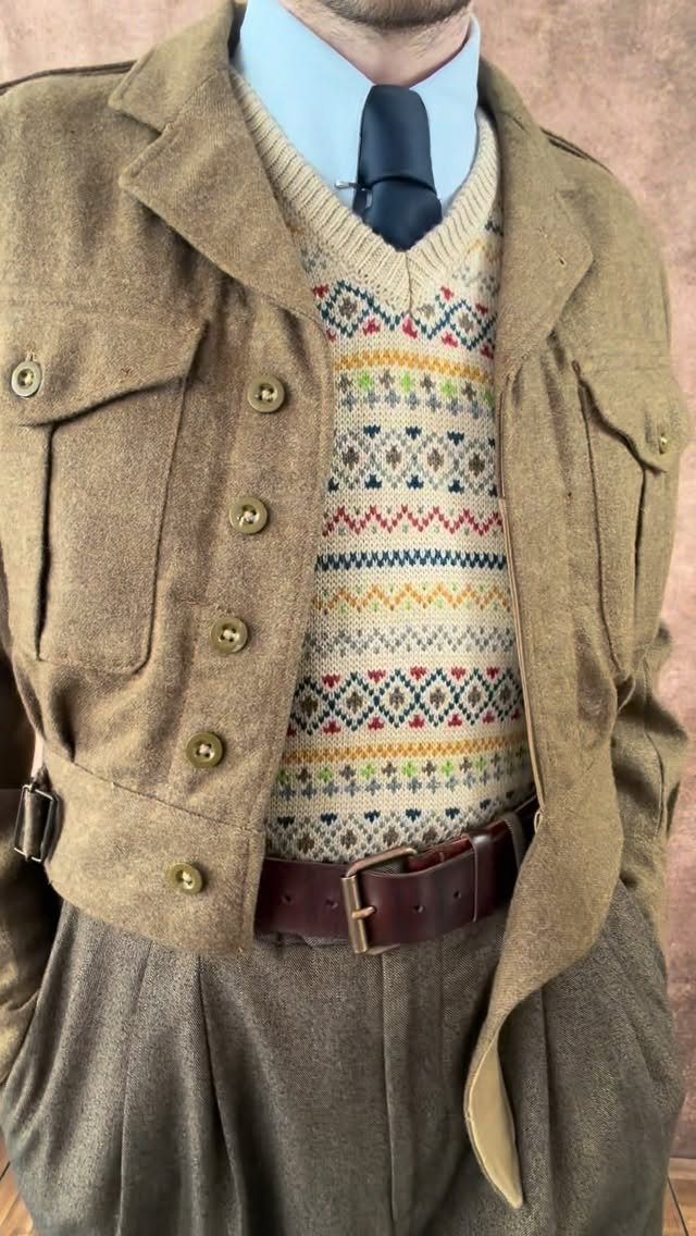 Man wearing a tan military jacket, patterned sweater vest, blue shirt, and dark tie.
