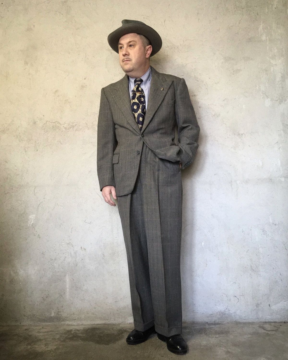 A man in a grey patterned sack suit and fedora standing against a wall.