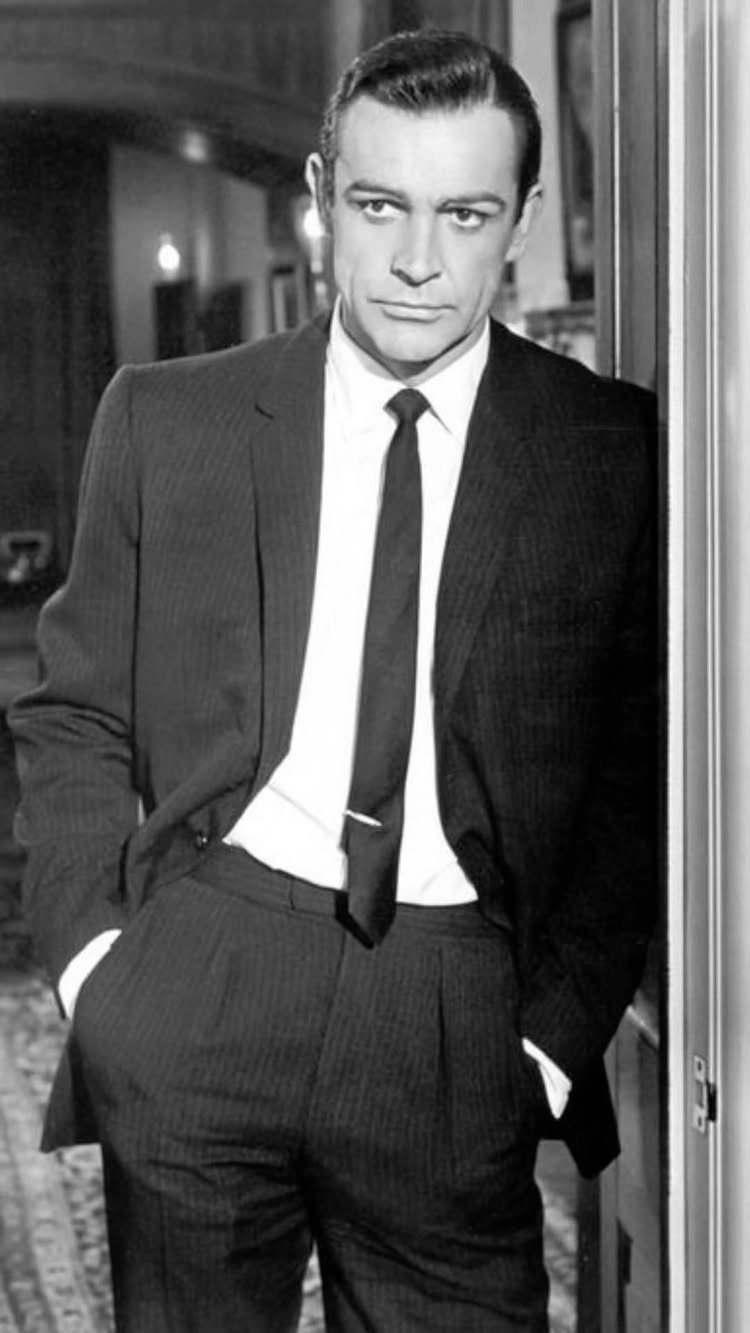 A man in a pinstripe suit and dark necktie leaning against a doorway.