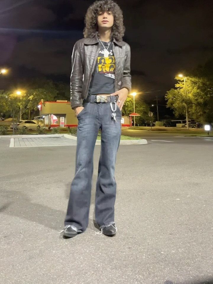 A person with curly hair wearing a leather jacket and flared jeans standing in a parking lot.