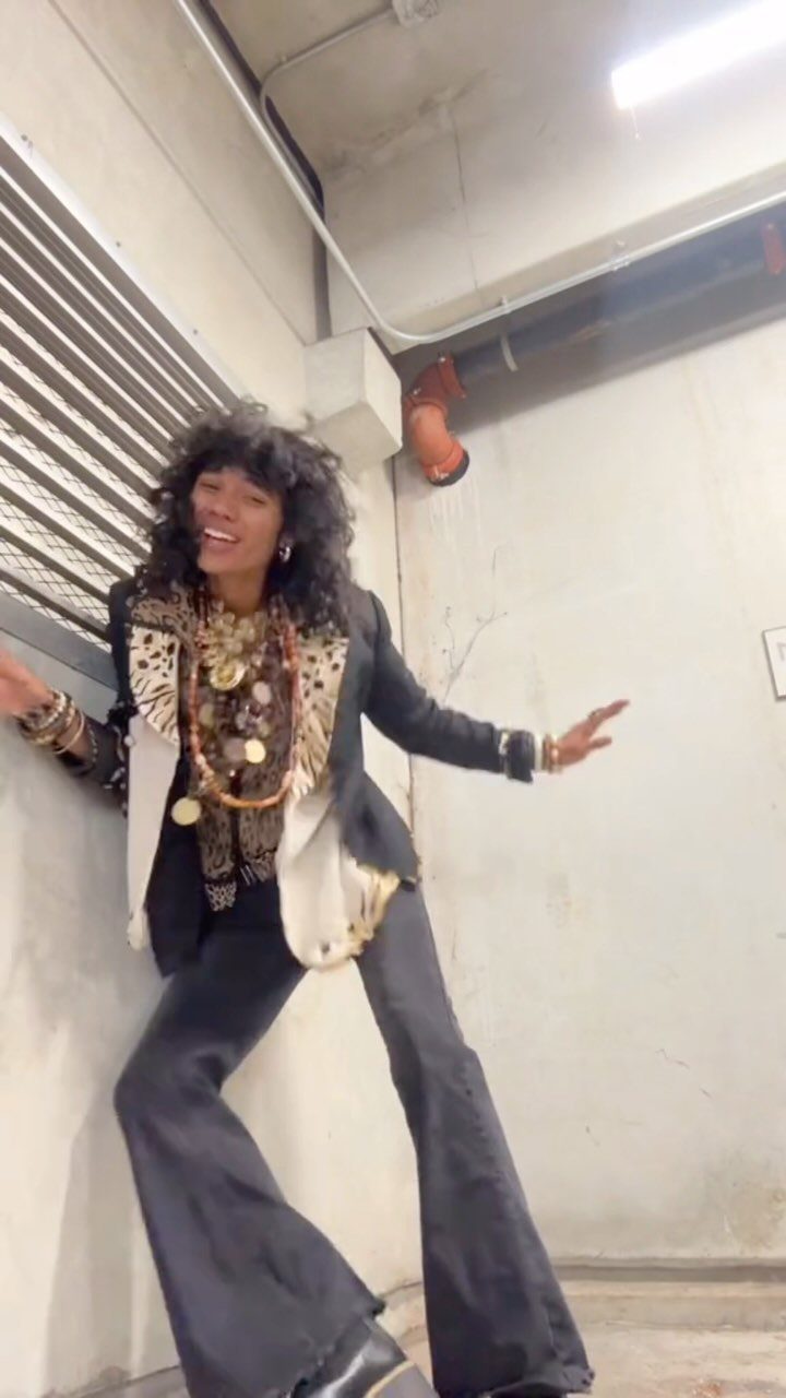 A person with curly hair posing in a patterned top, vest, and flared trousers with large gold necklaces.
