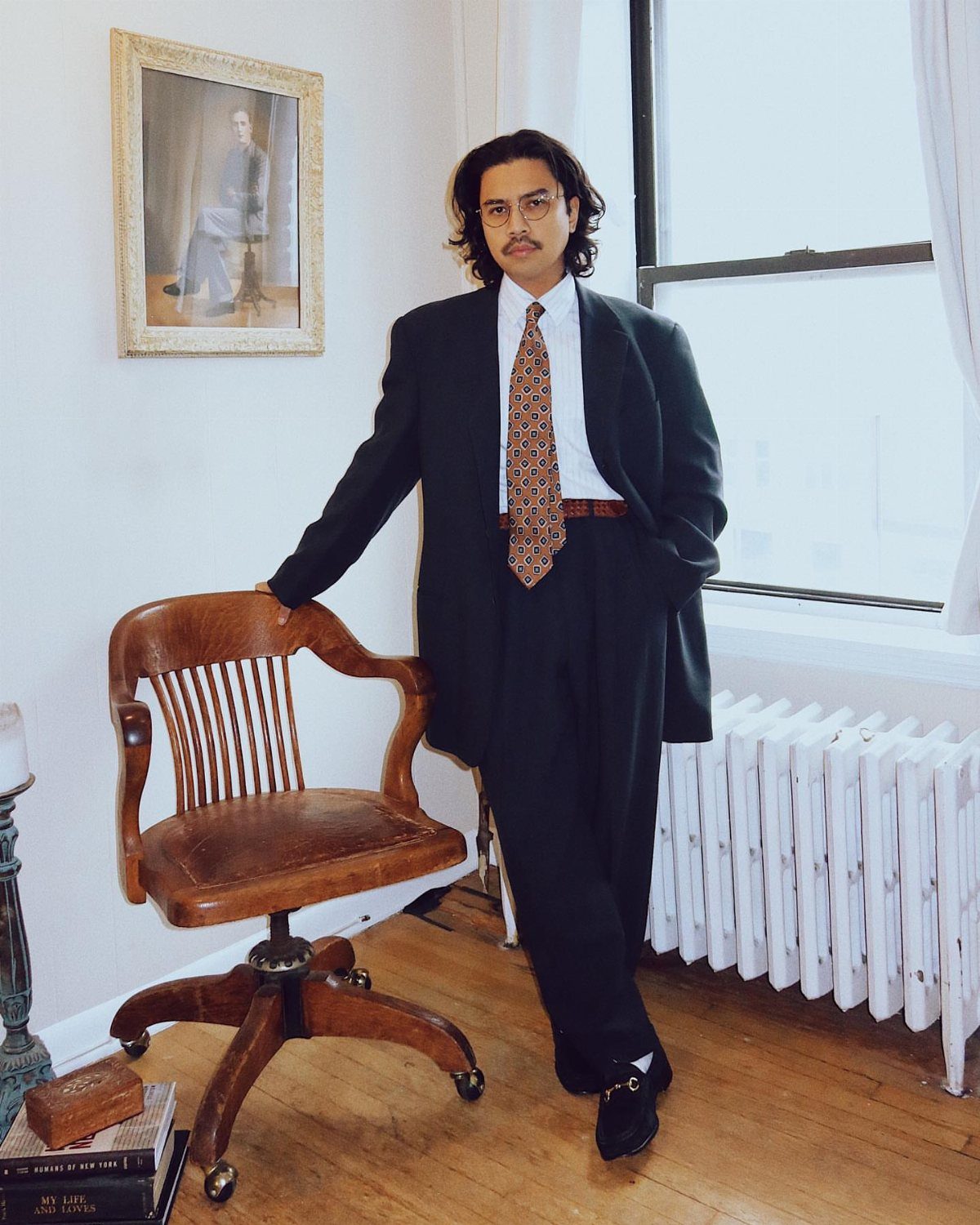 Man in a dark tailored suit and patterned tie standing next to a leather chair.