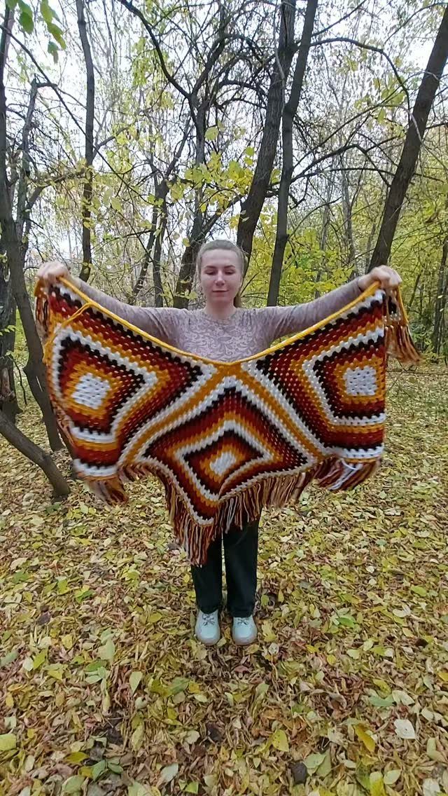 A woman holds up a large, colorful crochet cape with fringe in a forest.