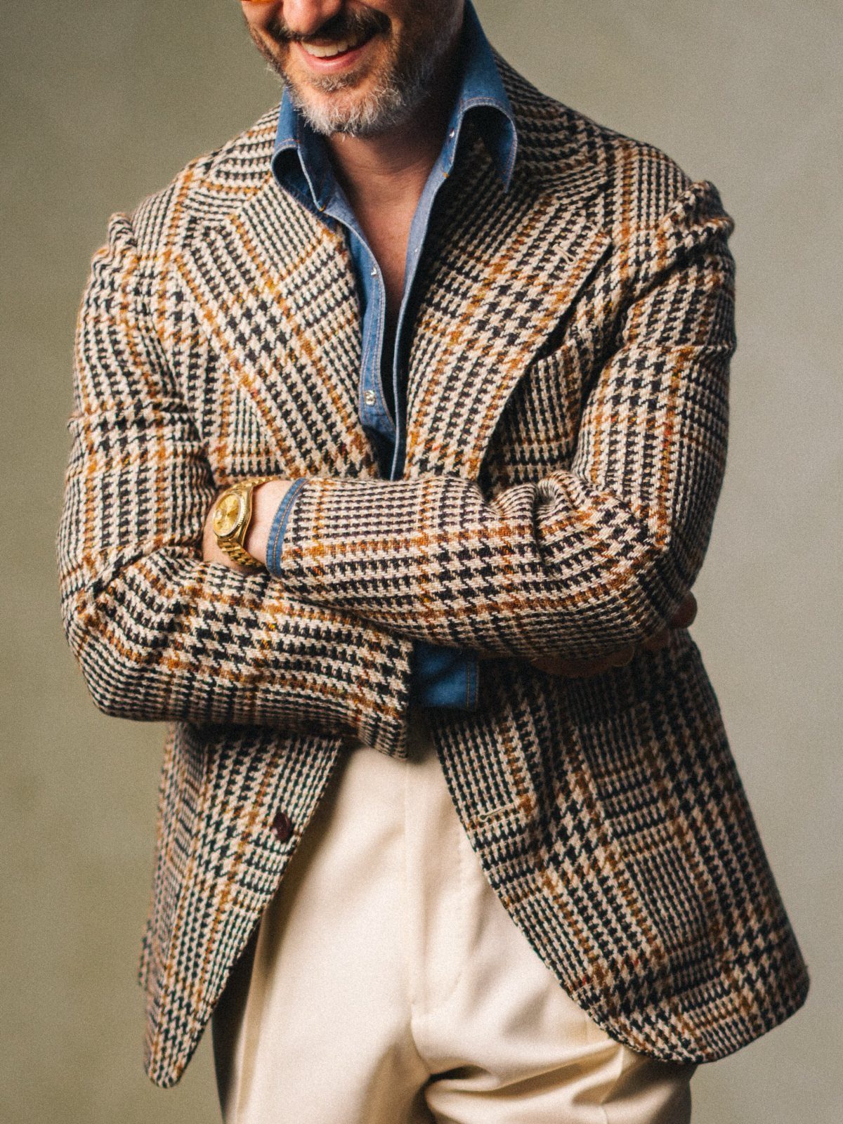 Man smiling in a patterned houndstooth blazer, blue denim shirt, and cream trousers.