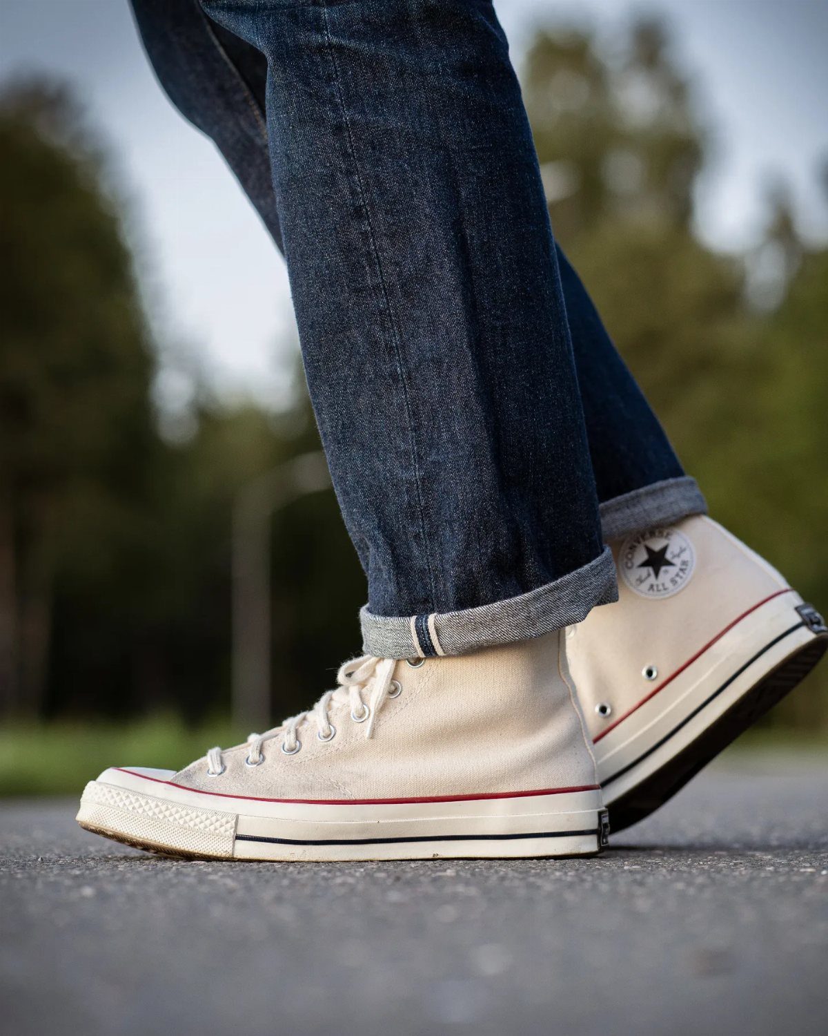 Cream high-top canvas sneakers worn with cuffed dark blue denim jeans on asphalt.