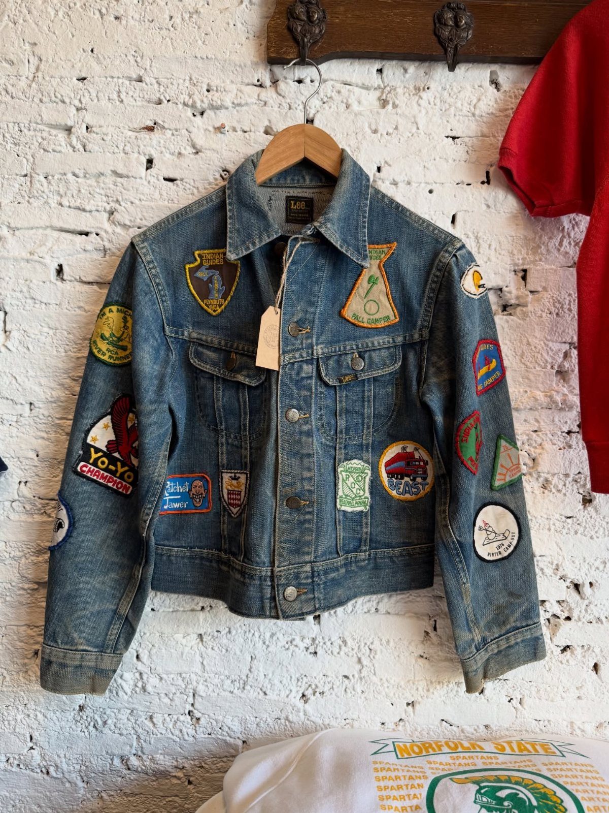 A blue denim jacket decorated with many colorful embroidered patches hanging on a white brick wall.