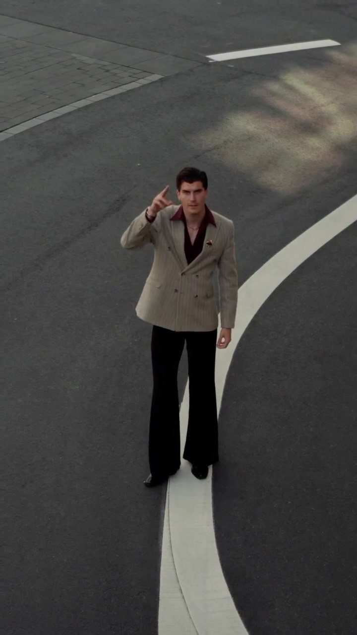 Man in a beige blazer and black flared trousers standing on a curved white road line.