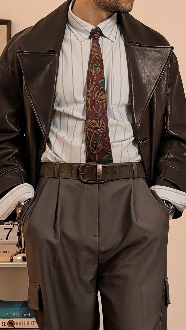 Man wearing a dark leather jacket, striped shirt, patterned tie, and grey pleated trousers.