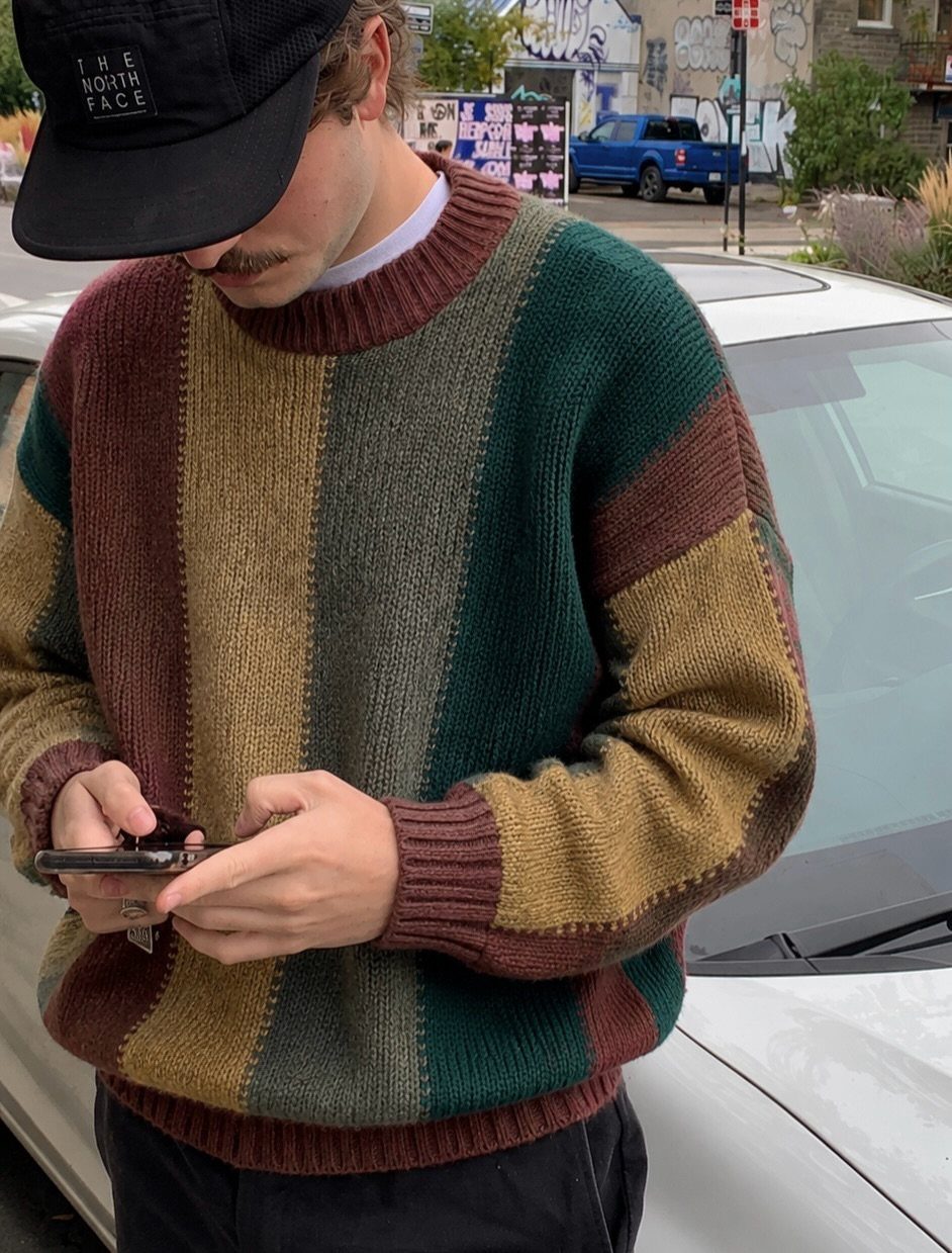 A man wearing a thick, multi-colored striped knit sweater while looking at his phone.