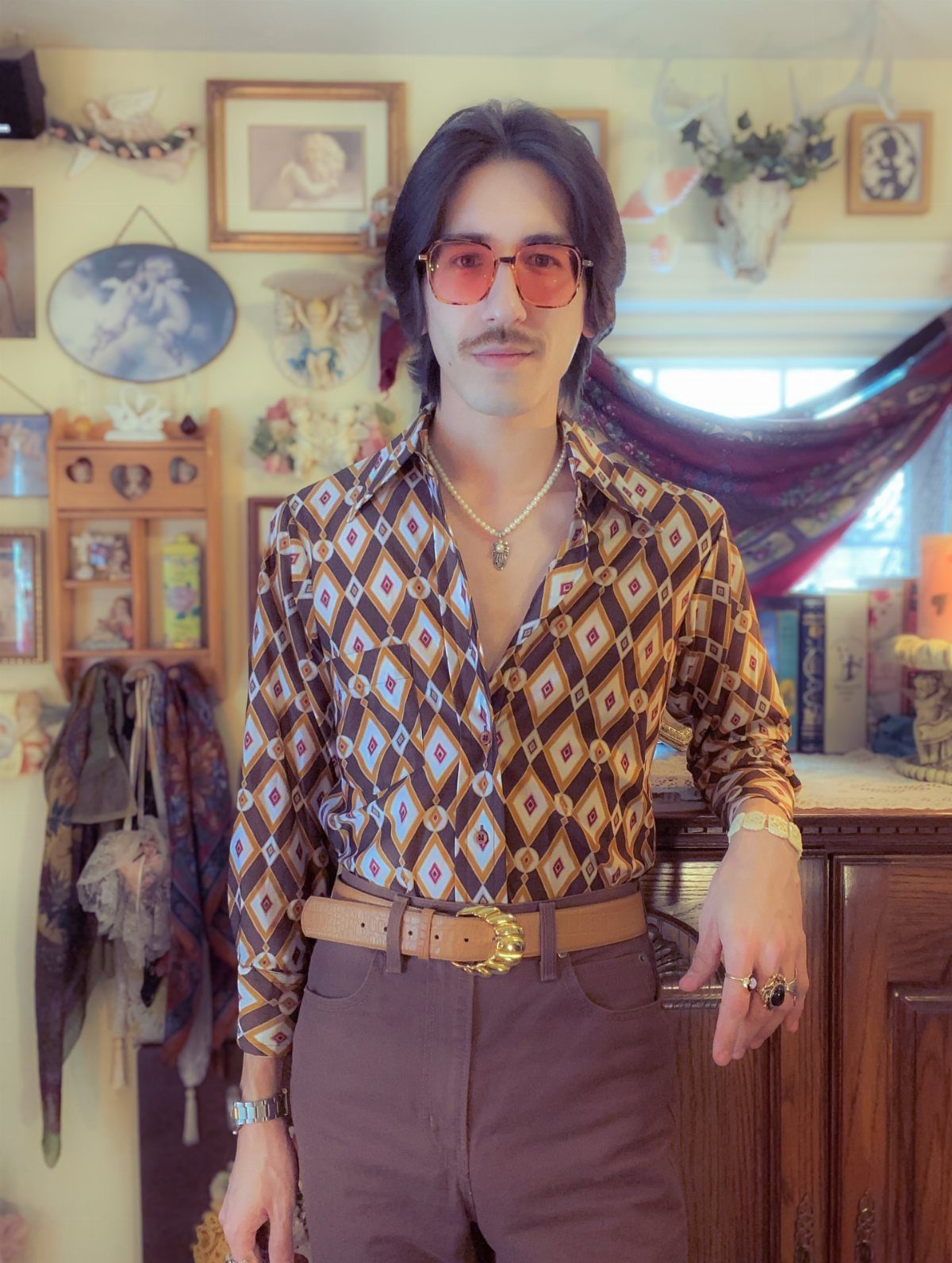 Man in a patterned brown shirt and tinted sunglasses standing in a vintage decorated room.