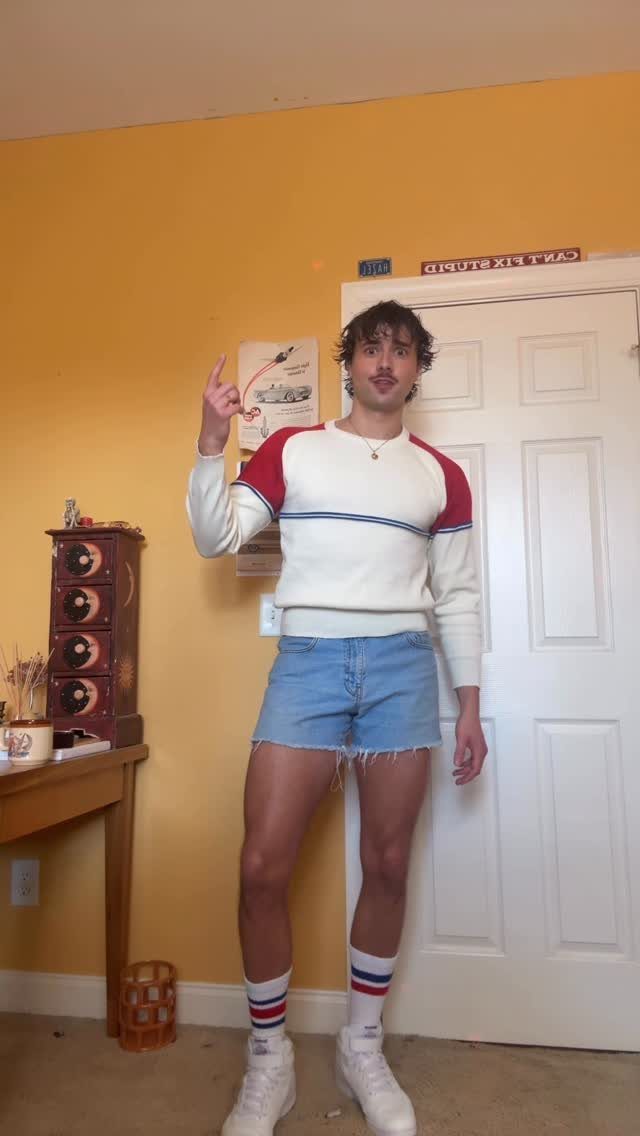 A young man posing in a white sweater and light blue denim short shorts.