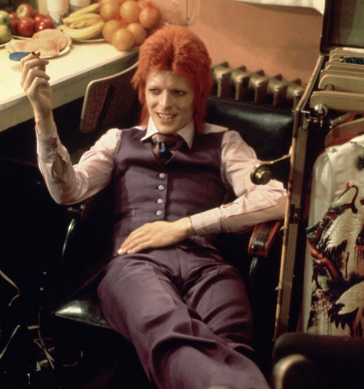 Red-haired person sitting in a chair wearing a purple waistcoat and matching trousers.