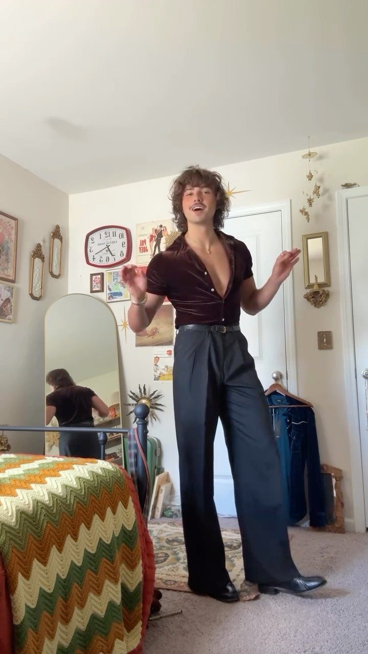 Man posing in a burgundy velvet shirt and black flared trousers in a vintage-style room.