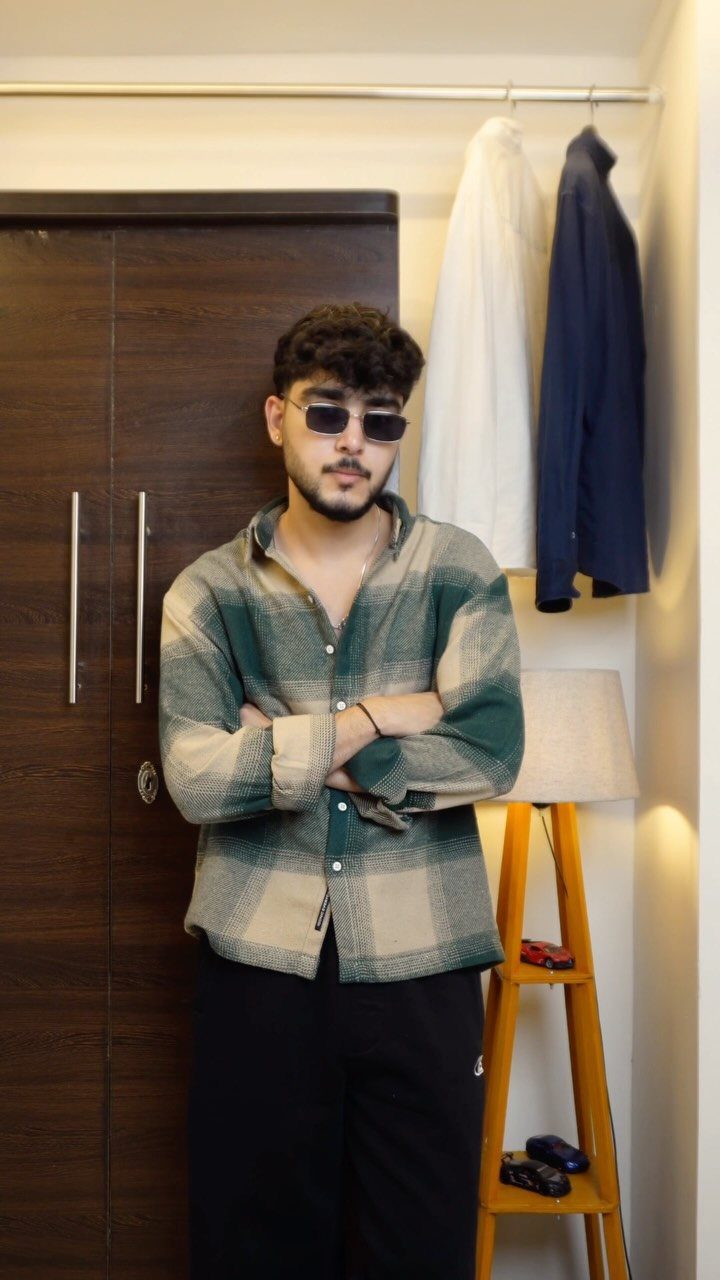 Man in plaid shirt and rectangular sunglasses posing indoors near a wardrobe and lamp.
