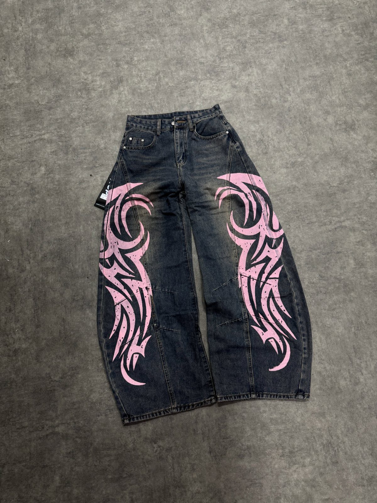 Dark denim baggy jeans with large pink tribal patterns laid on a grey surface.