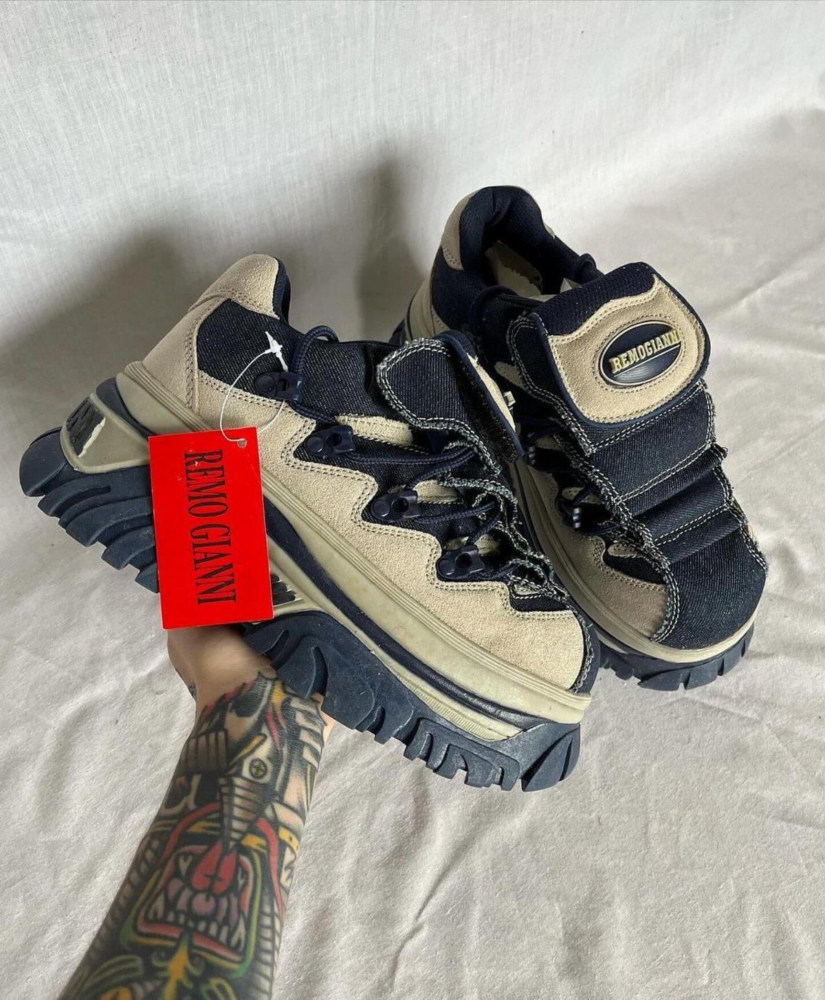 Chunky beige and navy blue sneakers with thick soles held by a tattooed arm.