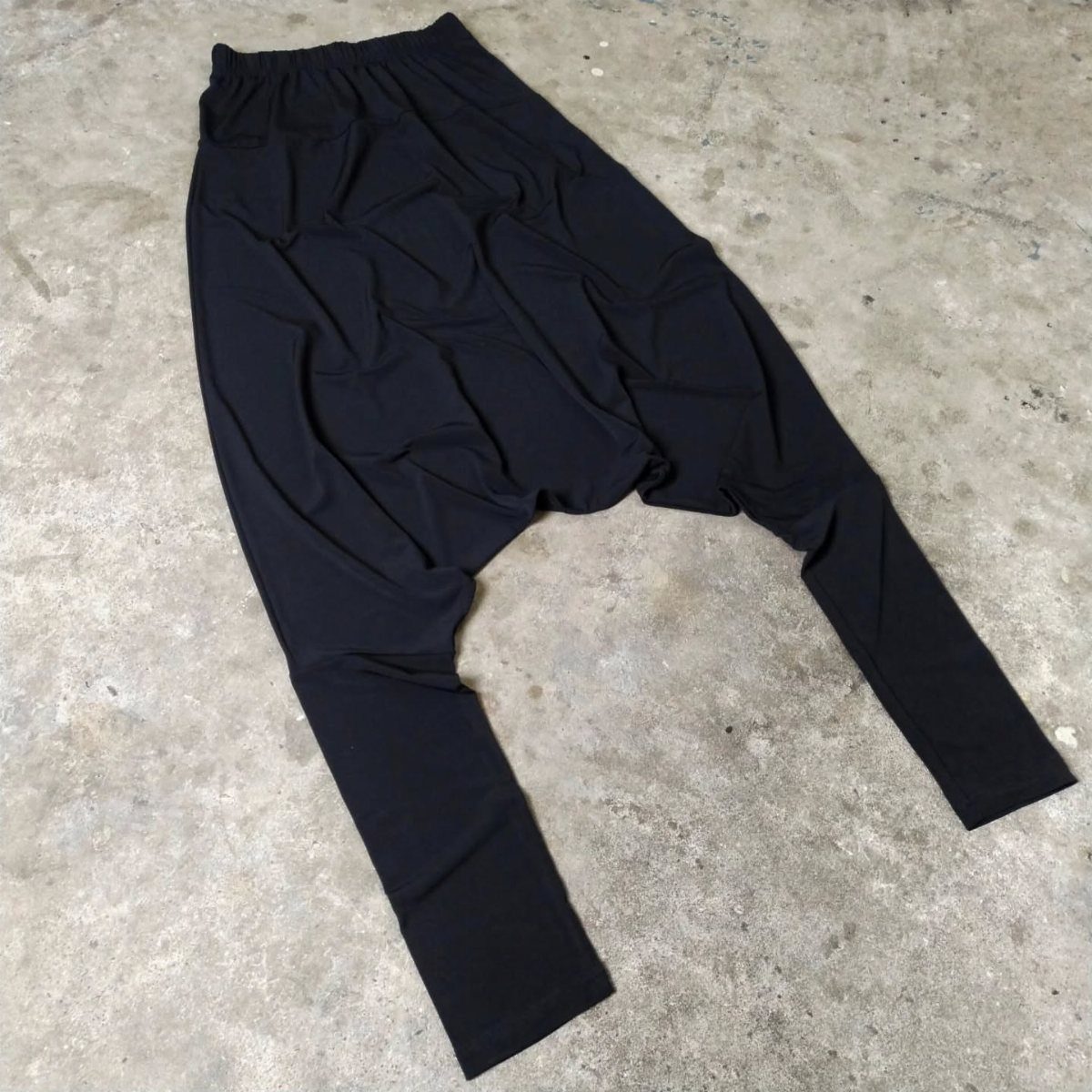 Black harem pants with a draped crotch laid flat on a grey concrete surface.