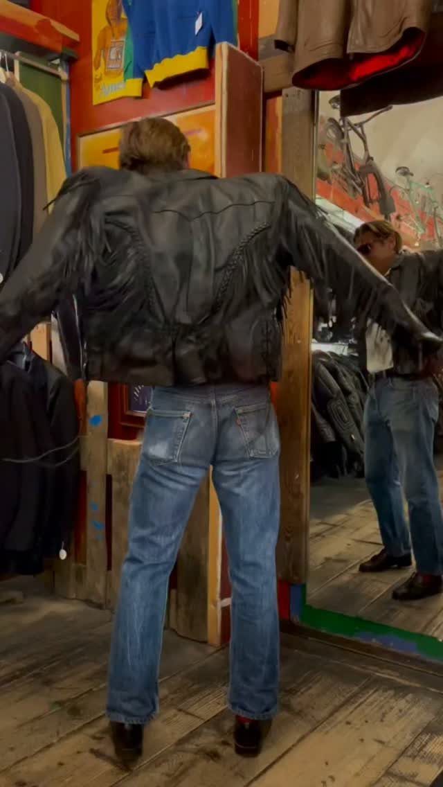 Man wearing a black leather fringe jacket and blue jeans standing in a clothing shop.