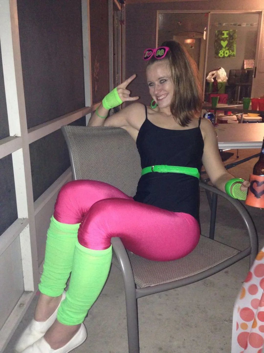Woman in bright pink leggings, black tank top, and neon green leg warmers sitting in a chair.