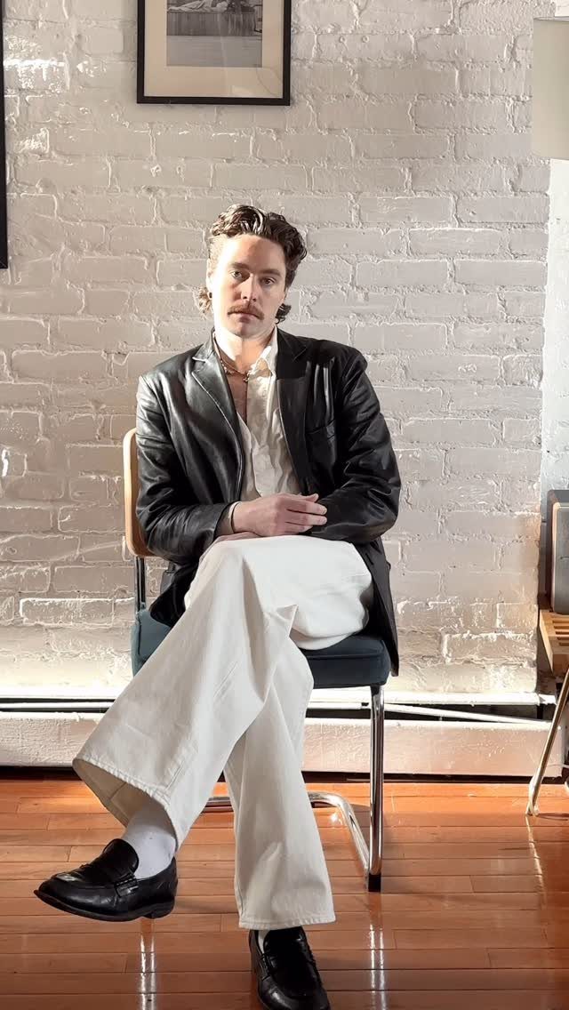 Man in a black leather blazer and wide-leg white trousers sitting on a chair.