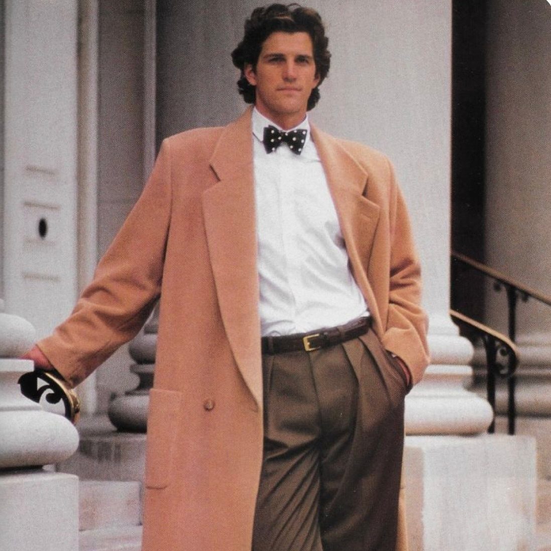 Man in a tan overcoat, white shirt, polka dot bowtie, and brown trousers standing by columns.