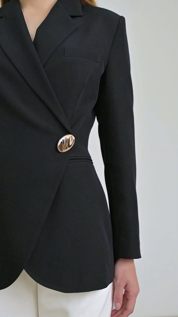 A black structured blazer with a large gold button worn over white trousers.
