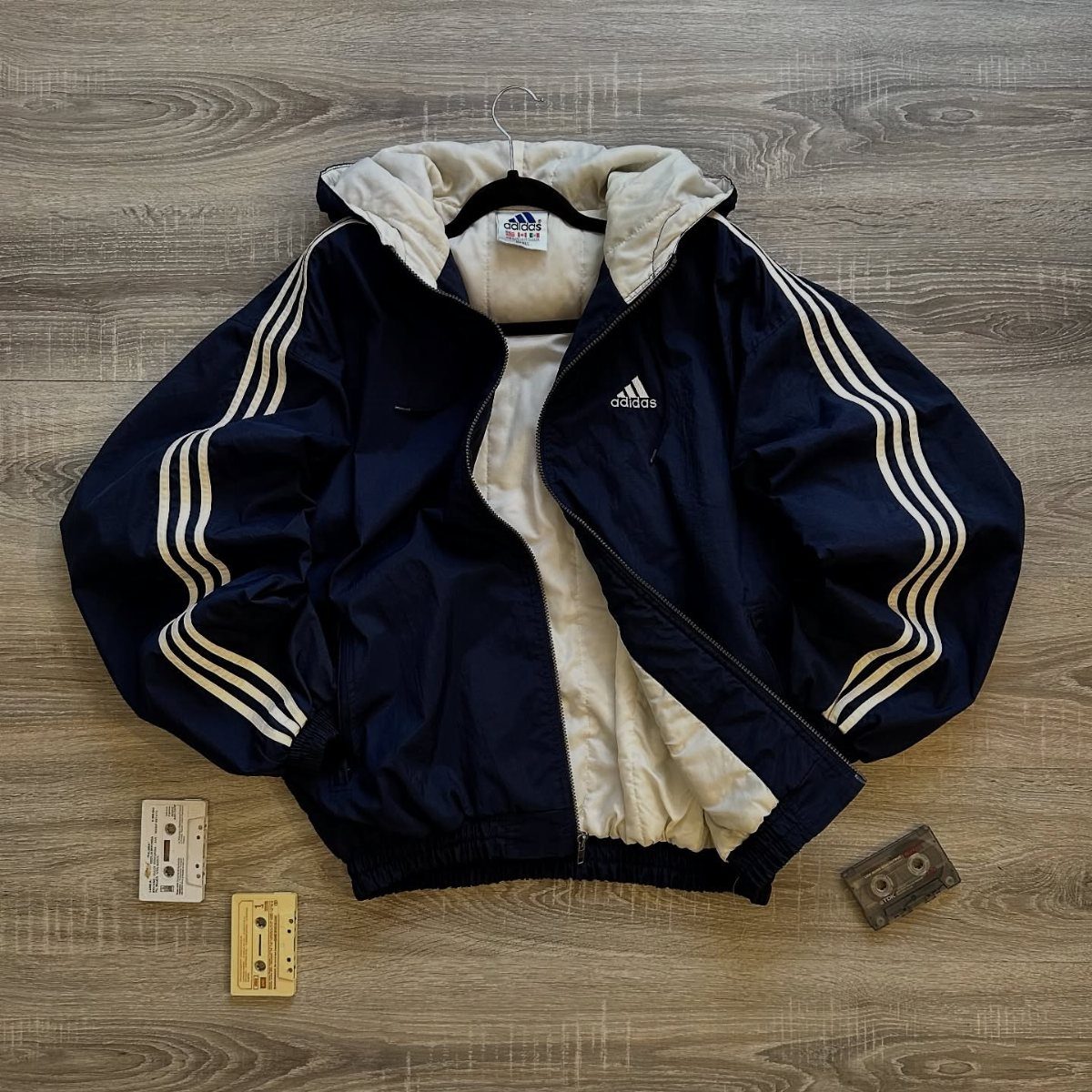 Navy blue Adidas windbreaker with white stripes and white lining on a wooden floor.