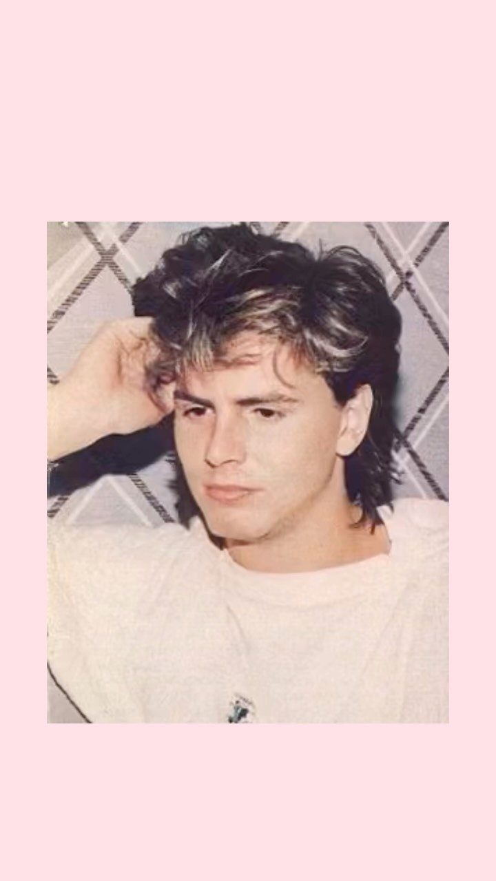 Young man with voluminous feathered hair posing against a plaid background in a white shirt.