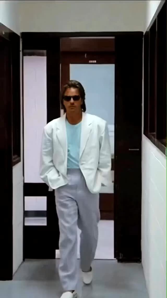 Man in a white blazer and light blue shirt walking through a doorway wearing sunglasses.
