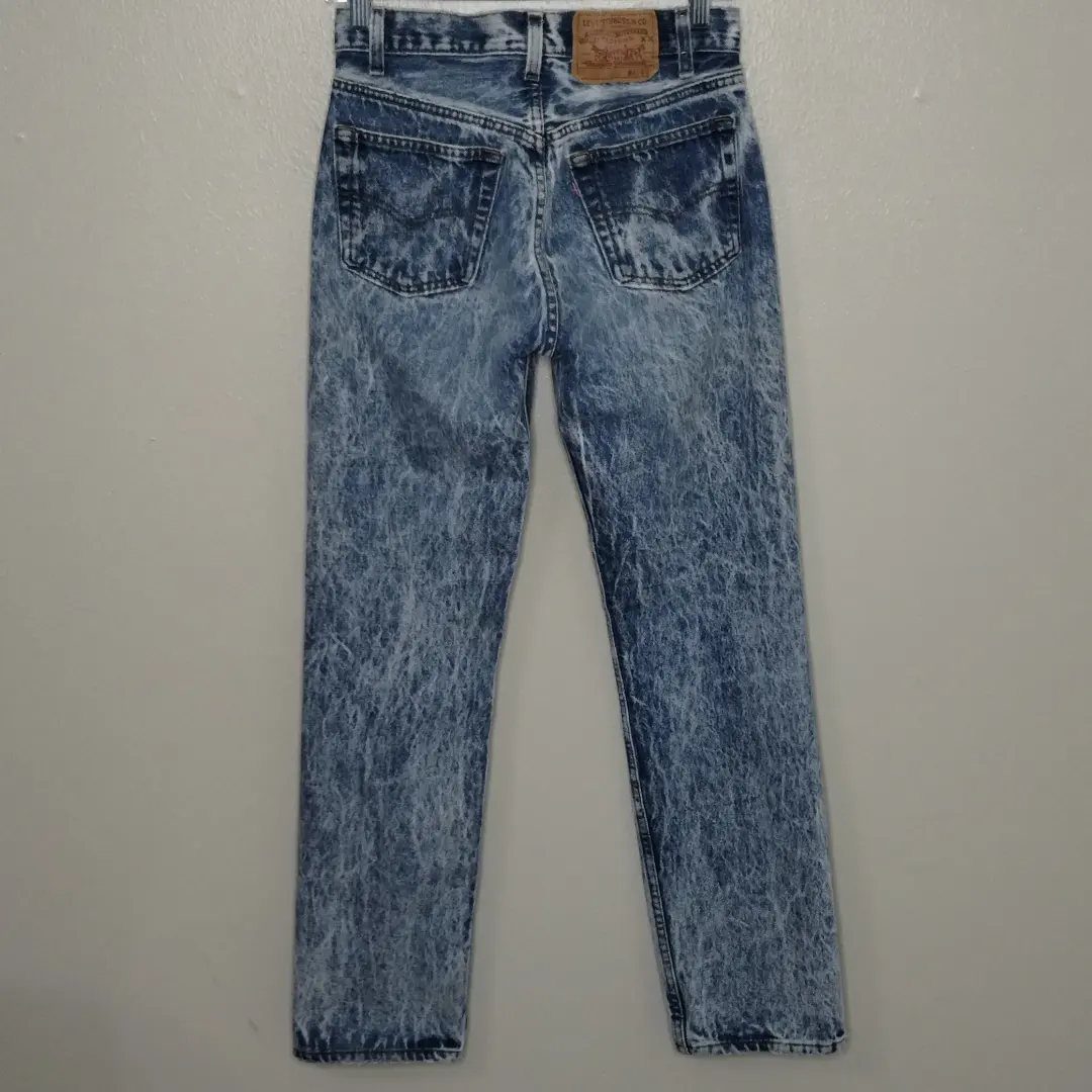 Acid-washed blue denim jeans with a mottled bleached pattern hanging against a grey wall.