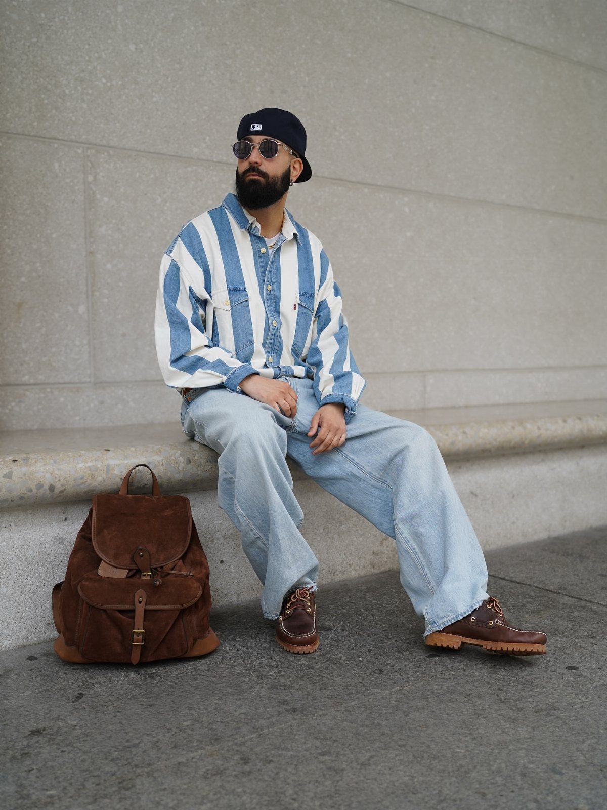A man wearing baggy denim jeans and a striped shirt sits on a concrete ledge.