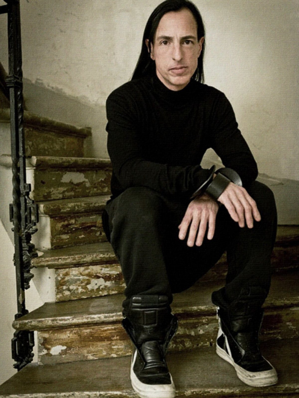 Man with long dark hair wearing black turtleneck and chunky sneakers sitting on wooden stairs.